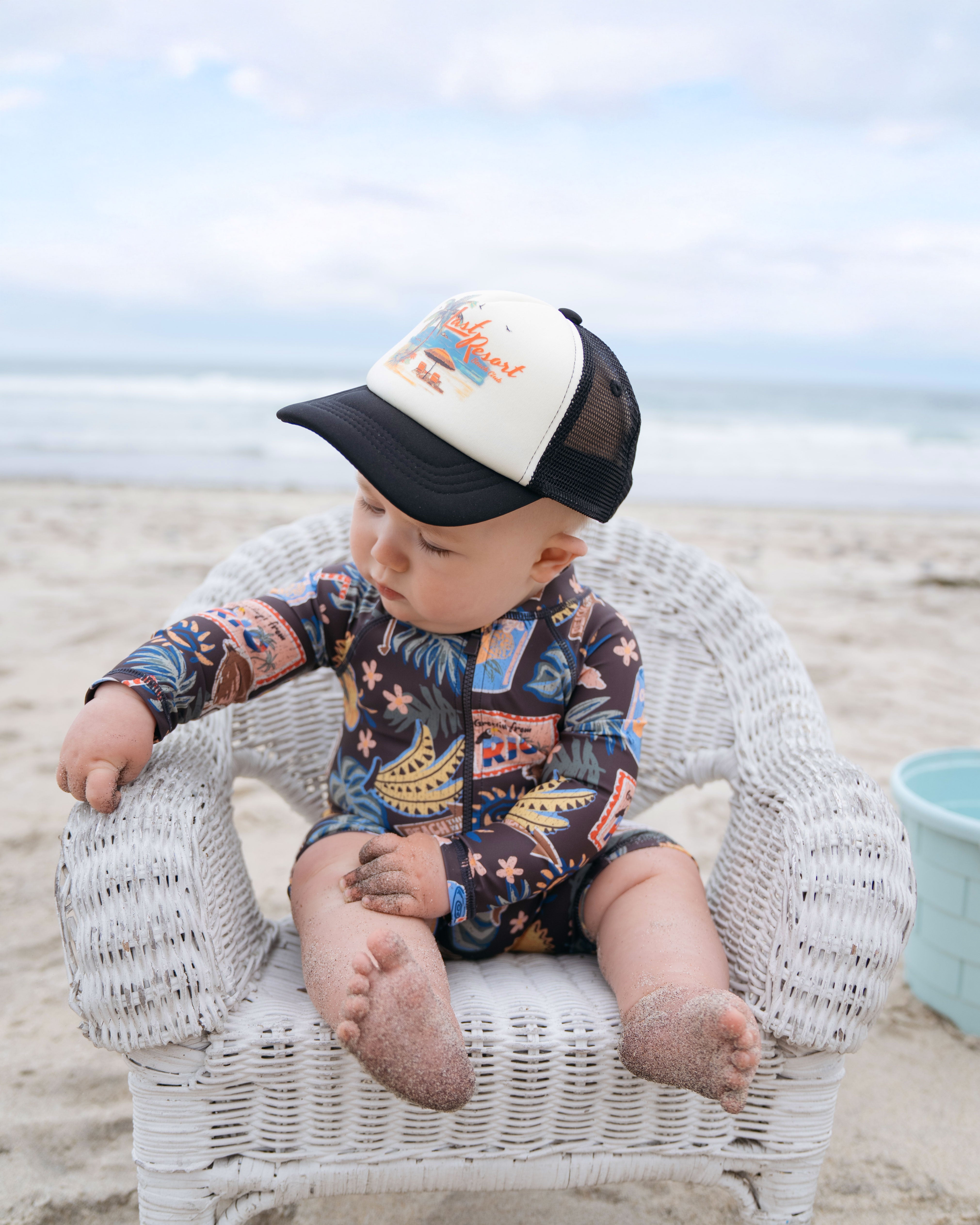 Baby sitting on a beach chair at the beach wearing a  Hemlock Foam trucker Ball Cap