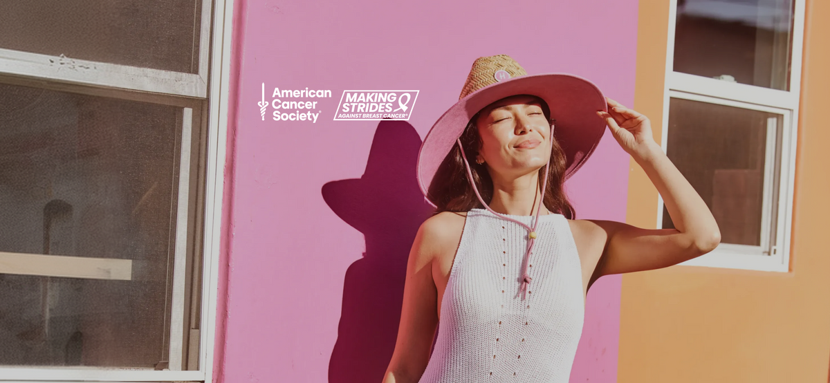 A woman wearing a white ribbed halter top and a wide-brimmed pink hat stands against a bright pink and orange wall, with the American Cancer Society Making Strides mark printed on the wall.