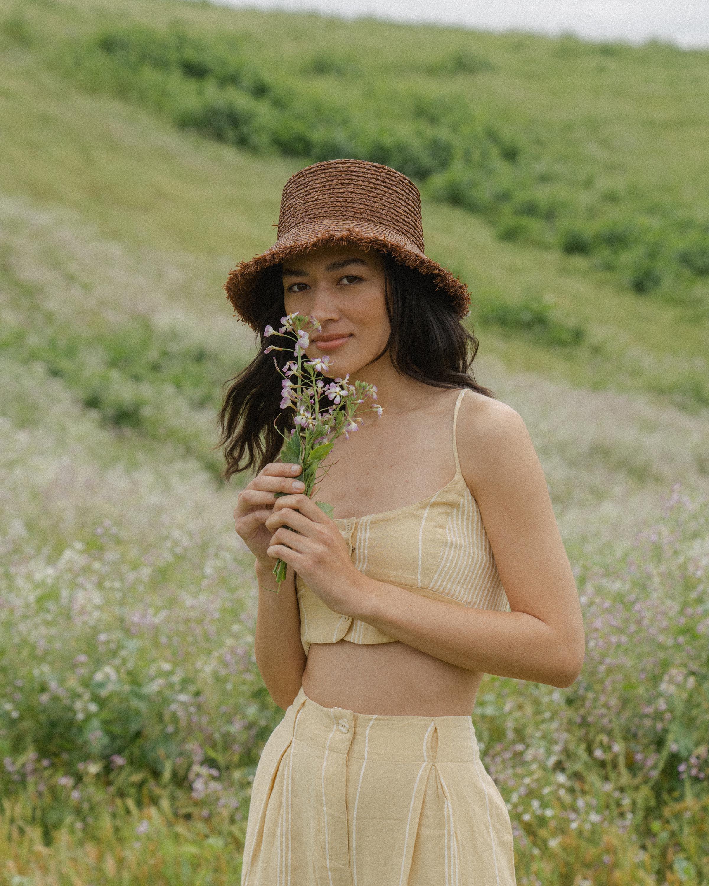 Hemlock female model looking straight wearing Lenny Straw Bucket Hat in Mocha holding a flower