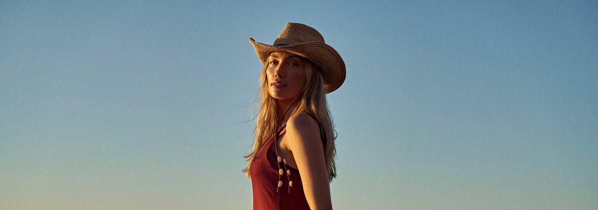 A woman wearing a straw cowboy hat and a flowing red dress stands on a beach at sunset, holding the hem of her dress as if to show off the fit or style.