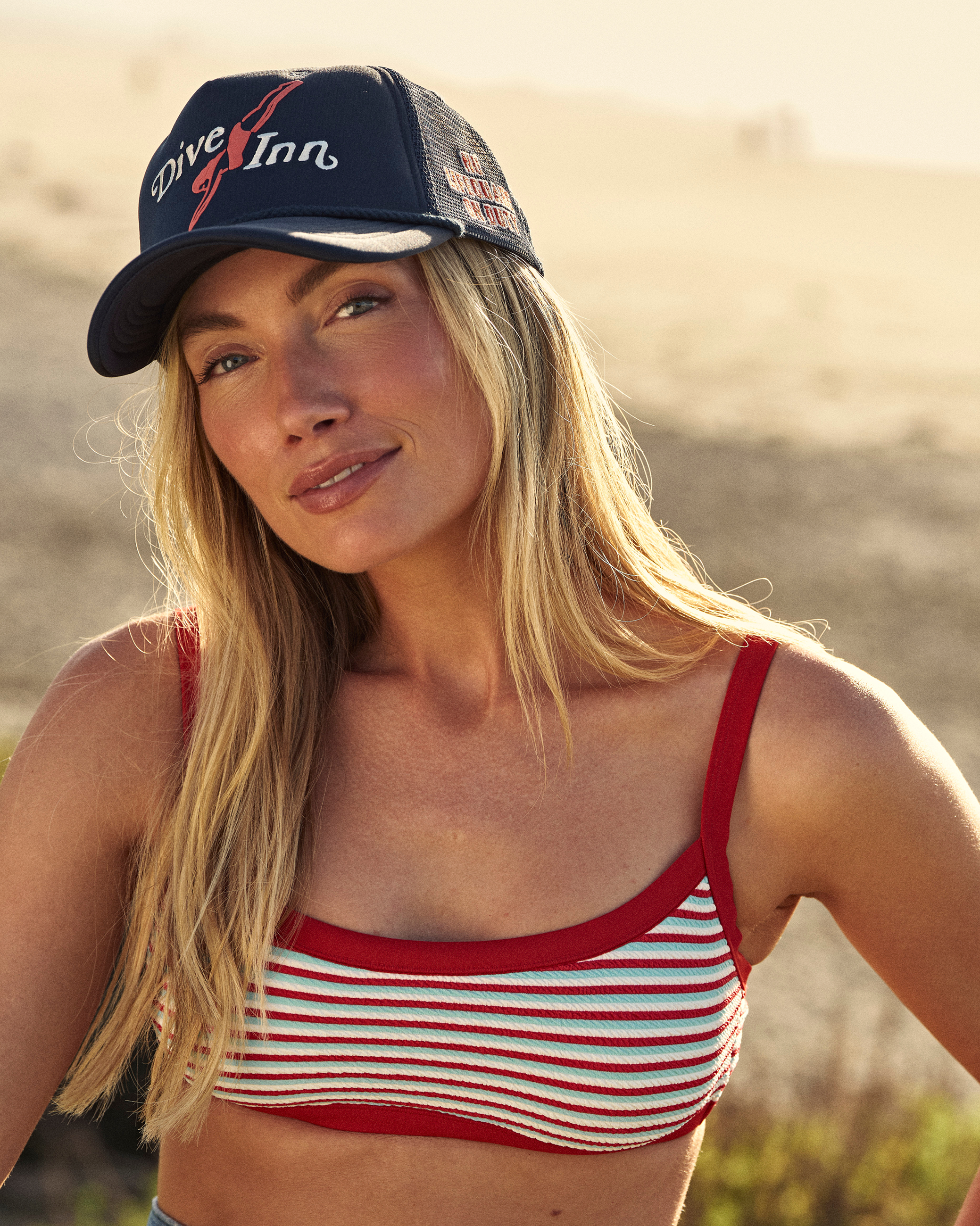Hemlock female model wearing the Navy blue Trucker Hat with 'Dive Inn' logo by the beach